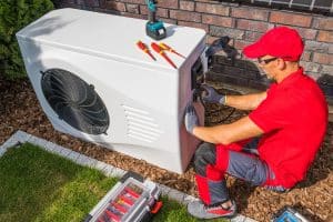 HVAC technician in a red uniform and safety gloves repairing an outdoor heat pump unit beside a brick wall, with screwdrivers, a power tool, and a toolbox nearby.
